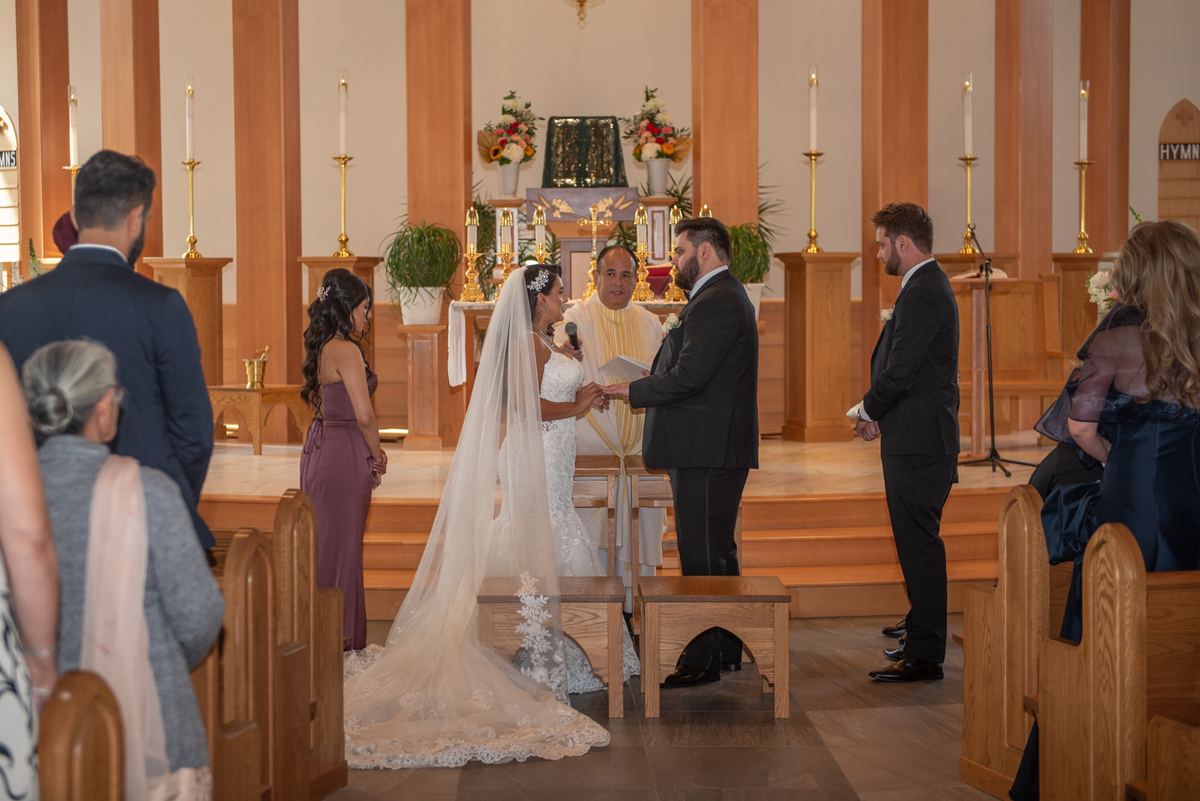 A bride and groom exchange vows at an altar, holding hands near a priest and floral arrangements in a wooden church.