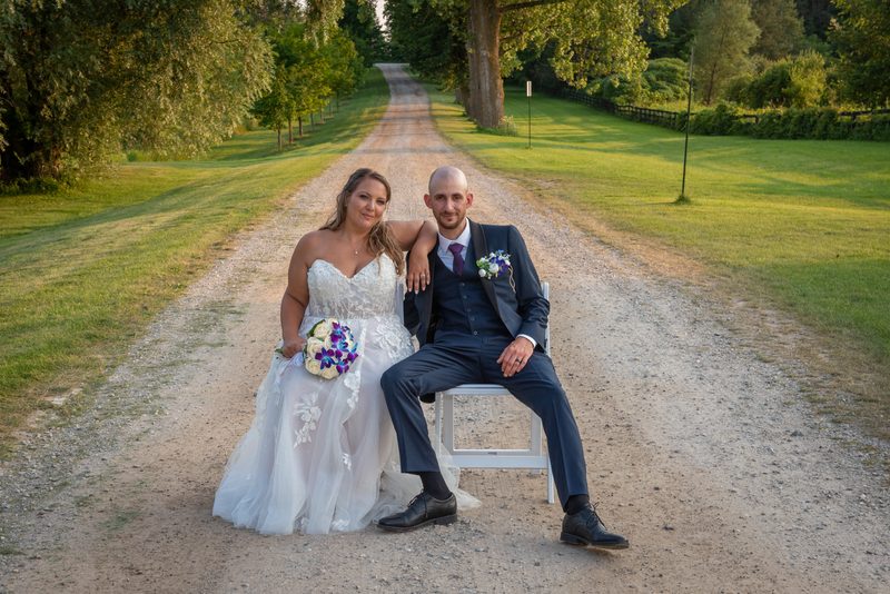 A newlywed couple in formal attire smiles on a sunlit gravel road, surrounded by trees and golden late afternoon light.