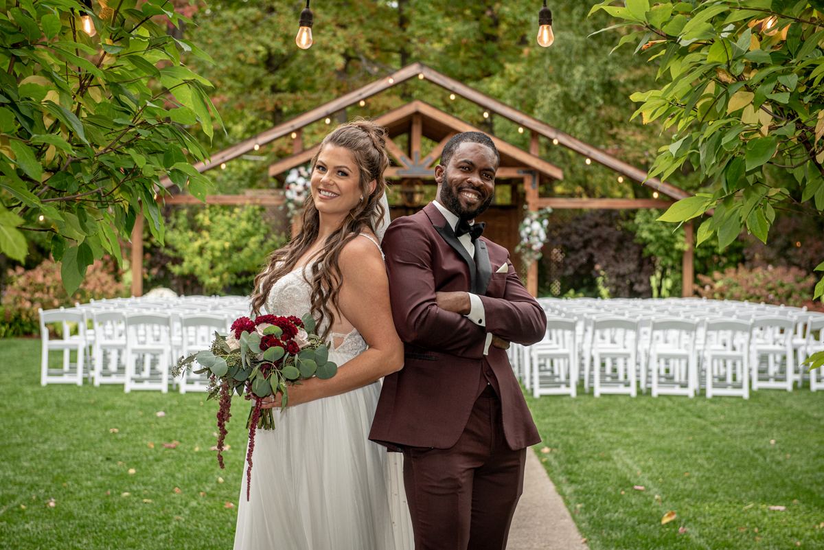 Bride and groom stand back-to-back smiling at camera in lush outdoor setting with string lights and greenery.