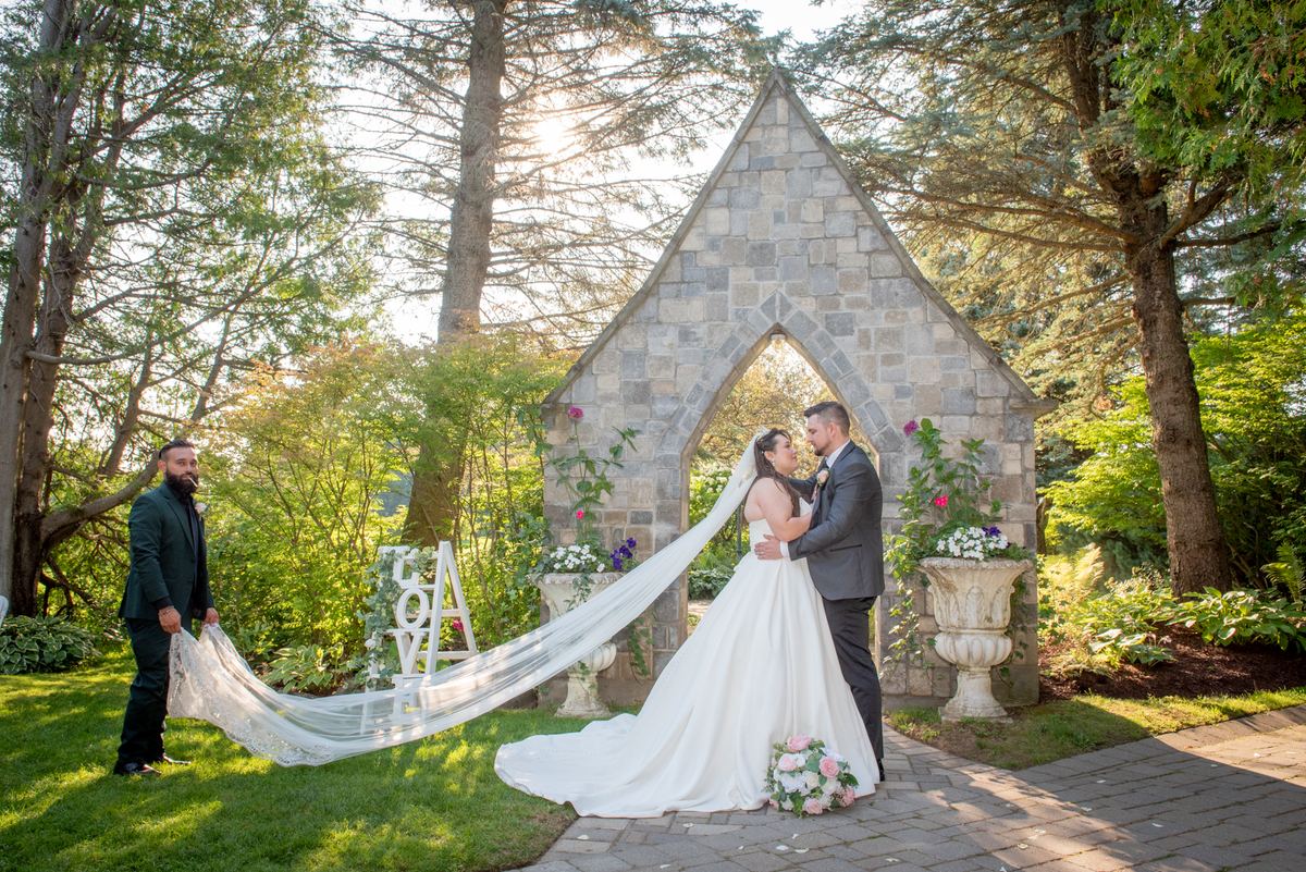 A bride and groom embrace under a stone arch in a sunlit garden, with a family member holding the veil.