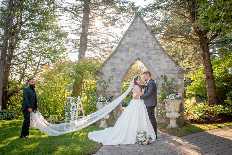 A bride and groom embrace under a stone arch in a sunlit garden, with a family member holding the veil.