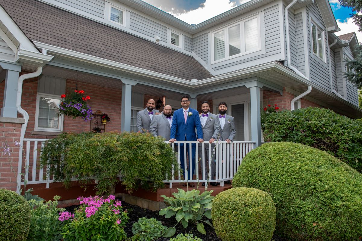 A groom in a blue suit stands with his gray-suited, purple-tied groomsmen on a sunny porch surrounded by green hedges.