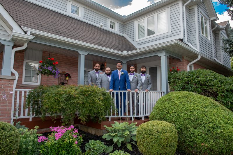 A groom in a blue suit stands with his gray-suited, purple-tied groomsmen on a sunny porch surrounded by green hedges.