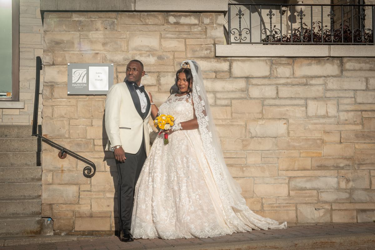 A bride in a lace gown and veil with yellow flowers stands beside her groom in a cream tuxedo near a stone wall.