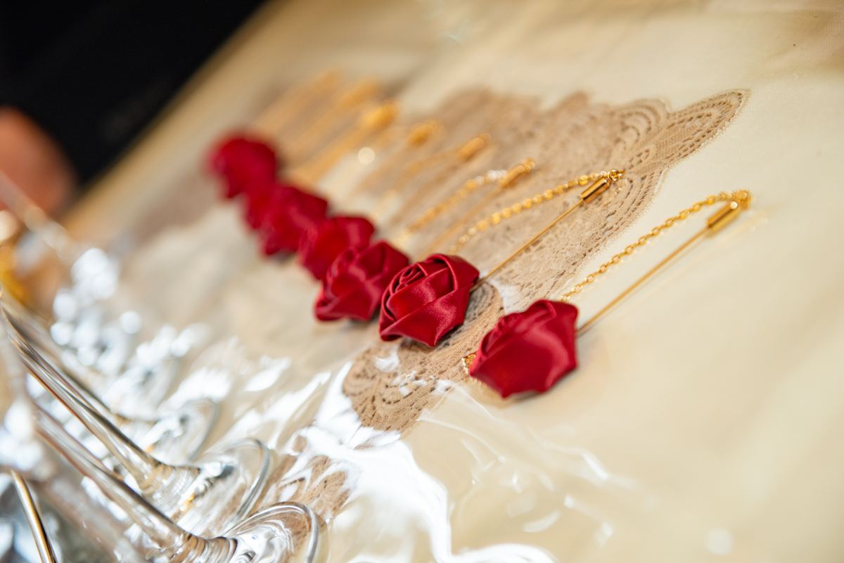 Red satin rose pins and gold chains lie on lace fabric beside glassware, evoking bridal preparation.