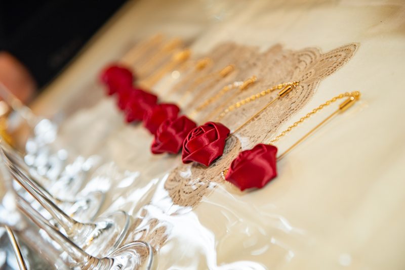 Red satin rose pins and gold chains lie on lace fabric beside glassware, evoking bridal preparation.