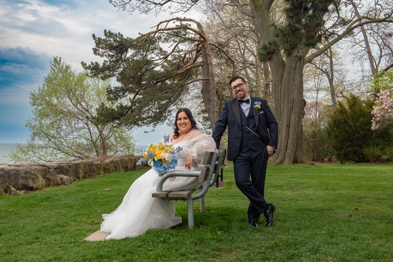 Selena and John share a smile near a waterfront, with Selena seated on a metal bench holding her bouquet of blue flowers.