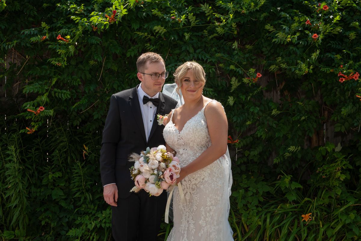 A bride in a lace gown and a groom in a tuxedo stand smiling in a garden filled with greenery and orange flowers.