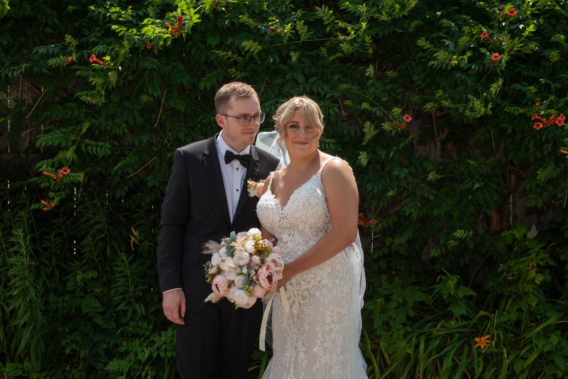 A bride in a lace gown and a groom in a tuxedo stand smiling in a garden filled with greenery and orange flowers.