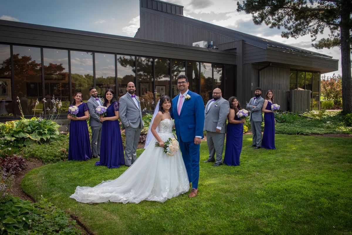A bride and groom stand with their bridal party in front of a modern building with large windows on a well-manicured lawn.