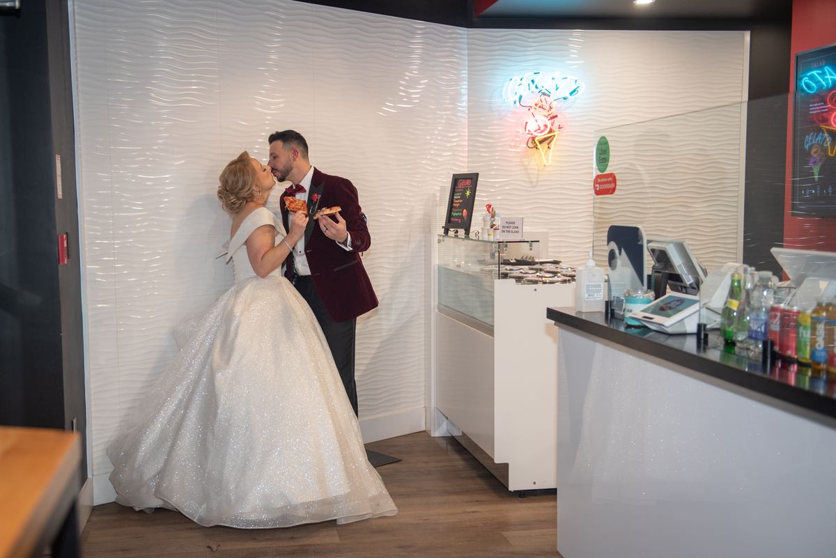 Bride and groom kiss while holding pizza slices in a dessert shop with warm lighting and neon sign, celebrating.
