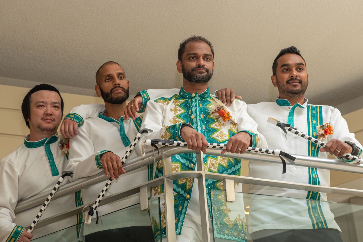 Four men in ceremonial attire stand on a balcony, holding decorated staffs and looking toward the camera.