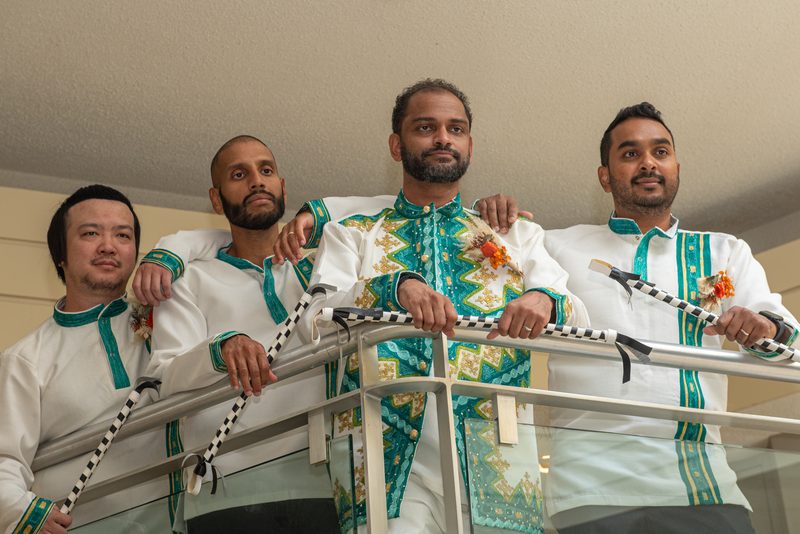 Four men in ceremonial attire stand on a balcony, holding decorated staffs and looking toward the camera.