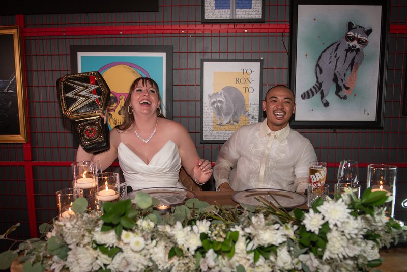 A bride and groom laugh beside a WWE belt at their decorated wedding table, with candles and framed art on the wall.