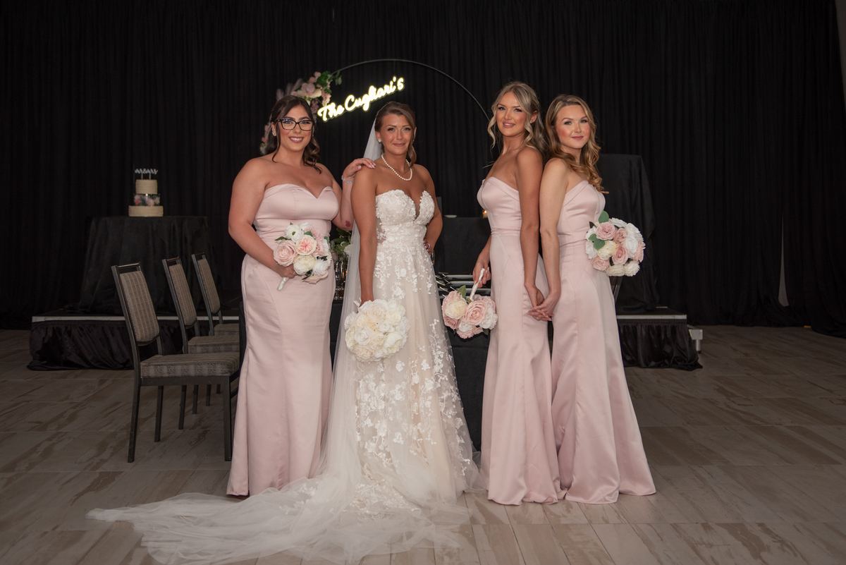 Bride in lace gown with bridesmaids in blush satin, smiling and holding flowers near a glowing sign in dim lighting.