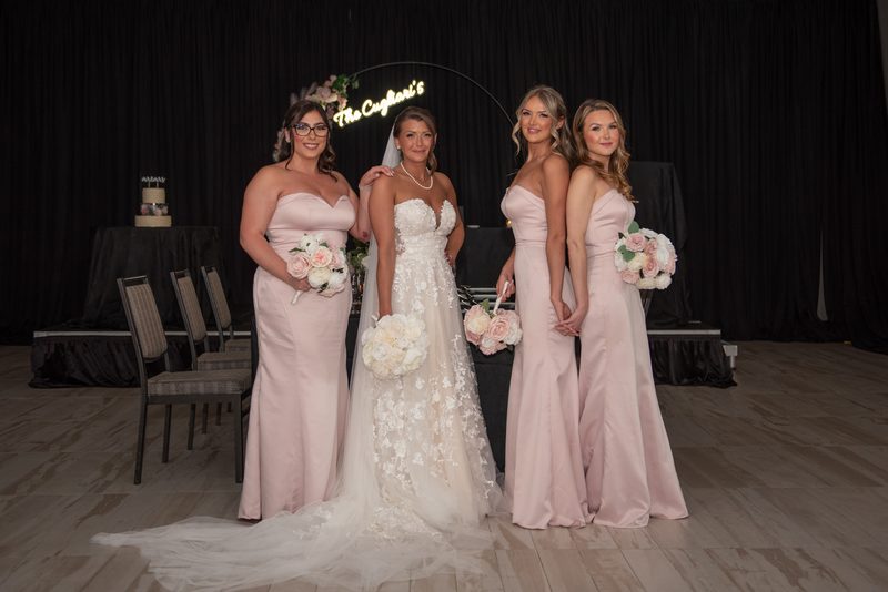 Bride in lace gown with bridesmaids in blush satin, smiling and holding flowers near a glowing sign in dim lighting.