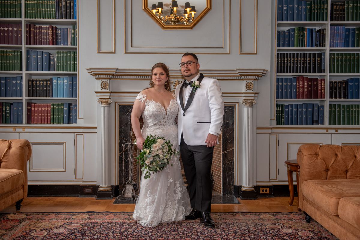 A bride and groom stand in a grand library with bookshelves lining the walls, smiling for the camera as rich wood floors.