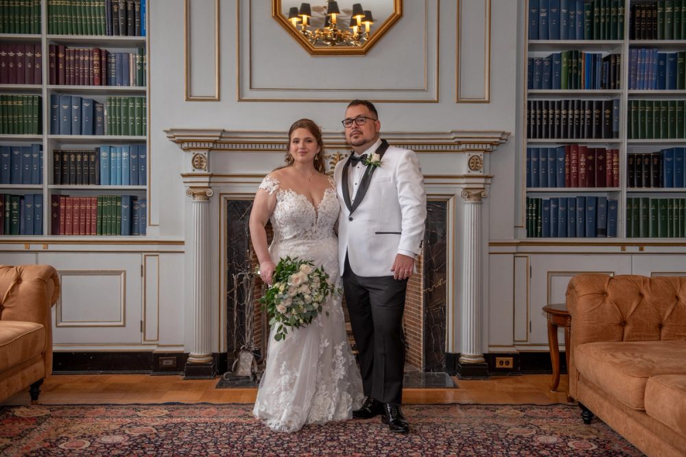 A bride and groom stand in a grand library with bookshelves lining the walls, smiling for the camera as rich wood floors.