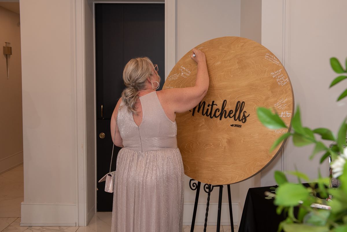 A woman in a wedding gown signs a wooden guestbook dedicated to Mitchells 2022, surrounded by indoor lighting.