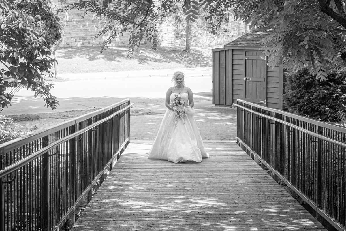 A bride in a white gown stands on a wooden bridge, smiling with a bouquet as sunlight filters through trees.