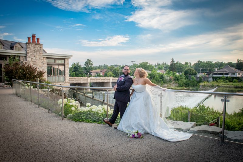 A newlywed couple poses by a river, the bride's gown trailing over the railing, in soft daylight with greenery.