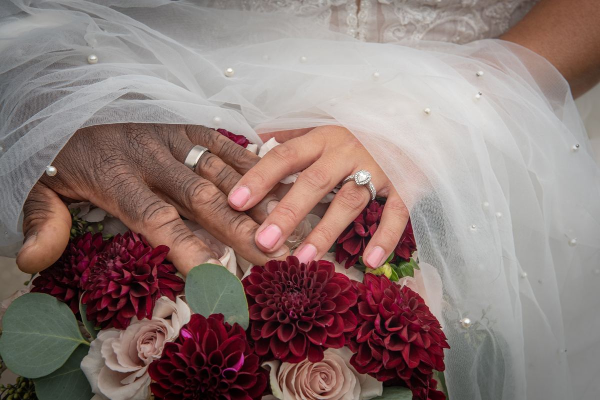 Two hands of different ages and skin tones overlap over a bouquet of dahlias and roses, veiled by tulle in soft light.