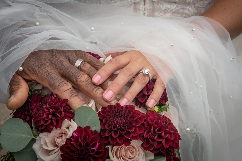 Two hands of different ages and skin tones overlap over a bouquet of dahlias and roses, veiled by tulle in soft light.
