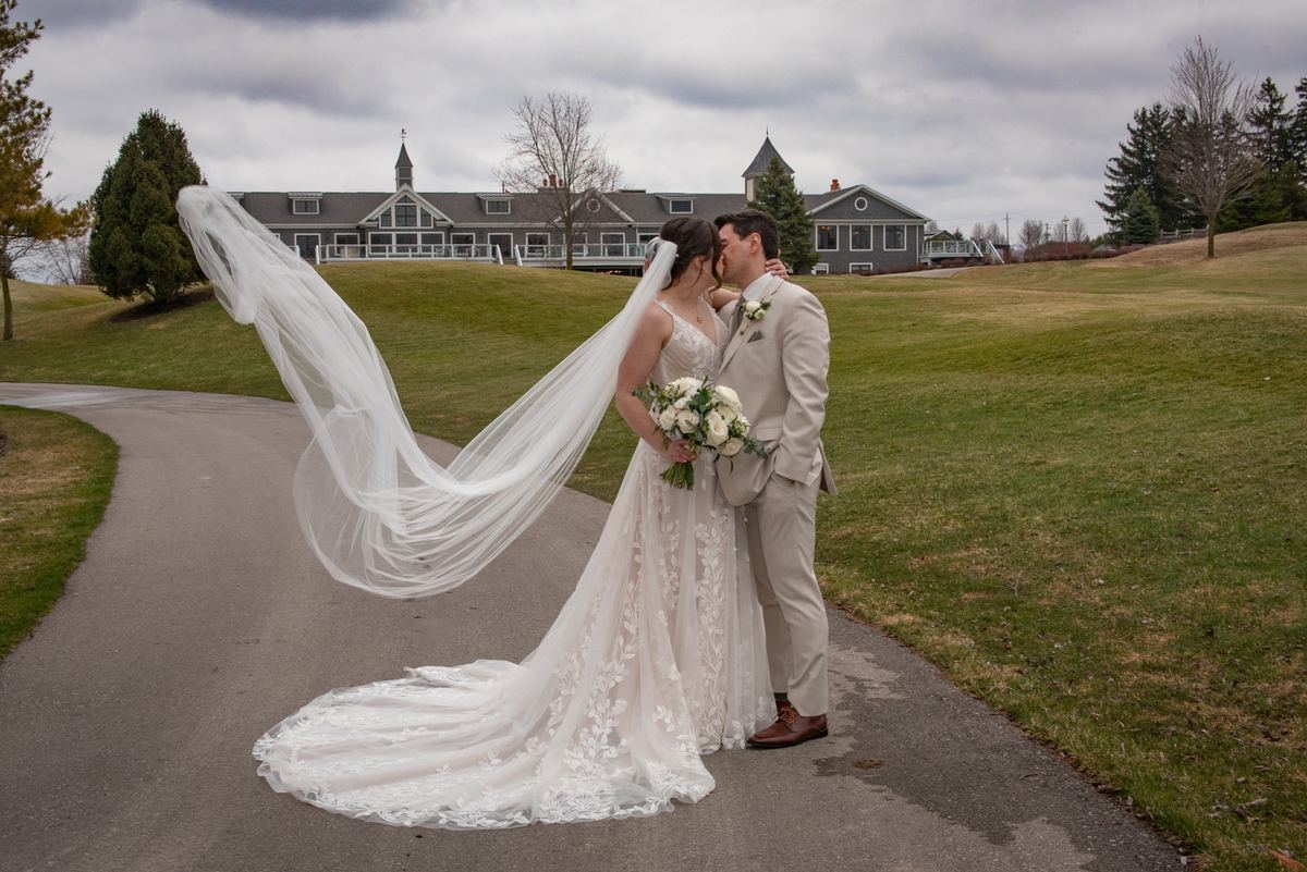 A bride and groom kiss along a path, her veil blowing in the wind over grassy land under an overcast sky.