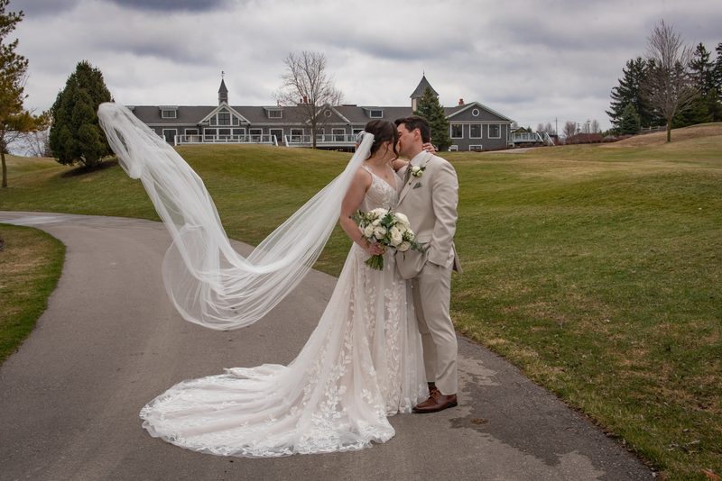 A bride and groom kiss along a path, her veil blowing in the wind over grassy land under an overcast sky.