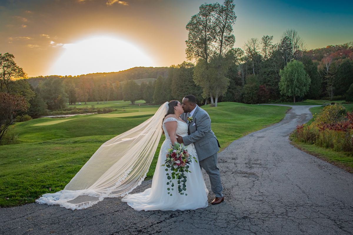 Bride and groom kiss beside a golf course path as the sun sets, casting warm light and long shadows.
