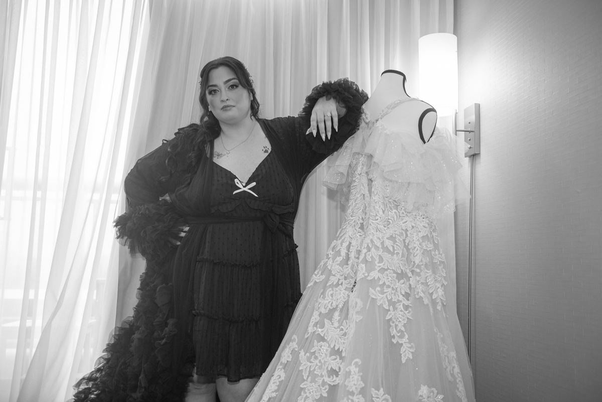 A woman in a dark robe stands beside a mannequin displaying a white wedding gown, with soft light filtering through curtains.