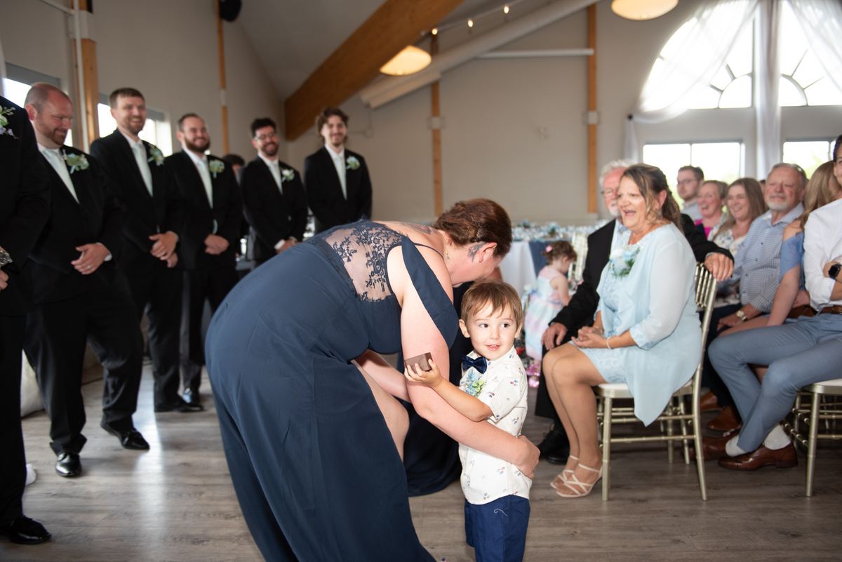 A woman in a navy wedding dress bends to embrace a young boy with a bow tie as guests smile and watch.