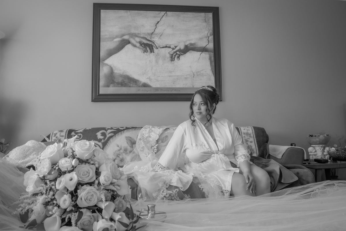 A bride sits thoughtfully on a patterned sofa with her bouquet and veil, with a reproduction of The Creation of Adam.