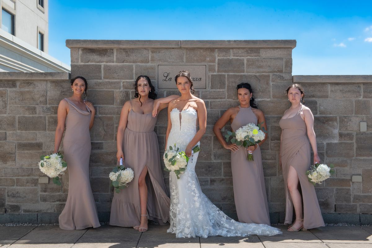 A bride in white lace and four bridesmaids in mauve dresses hold floral bouquets against a stone wall under clear blue skies.