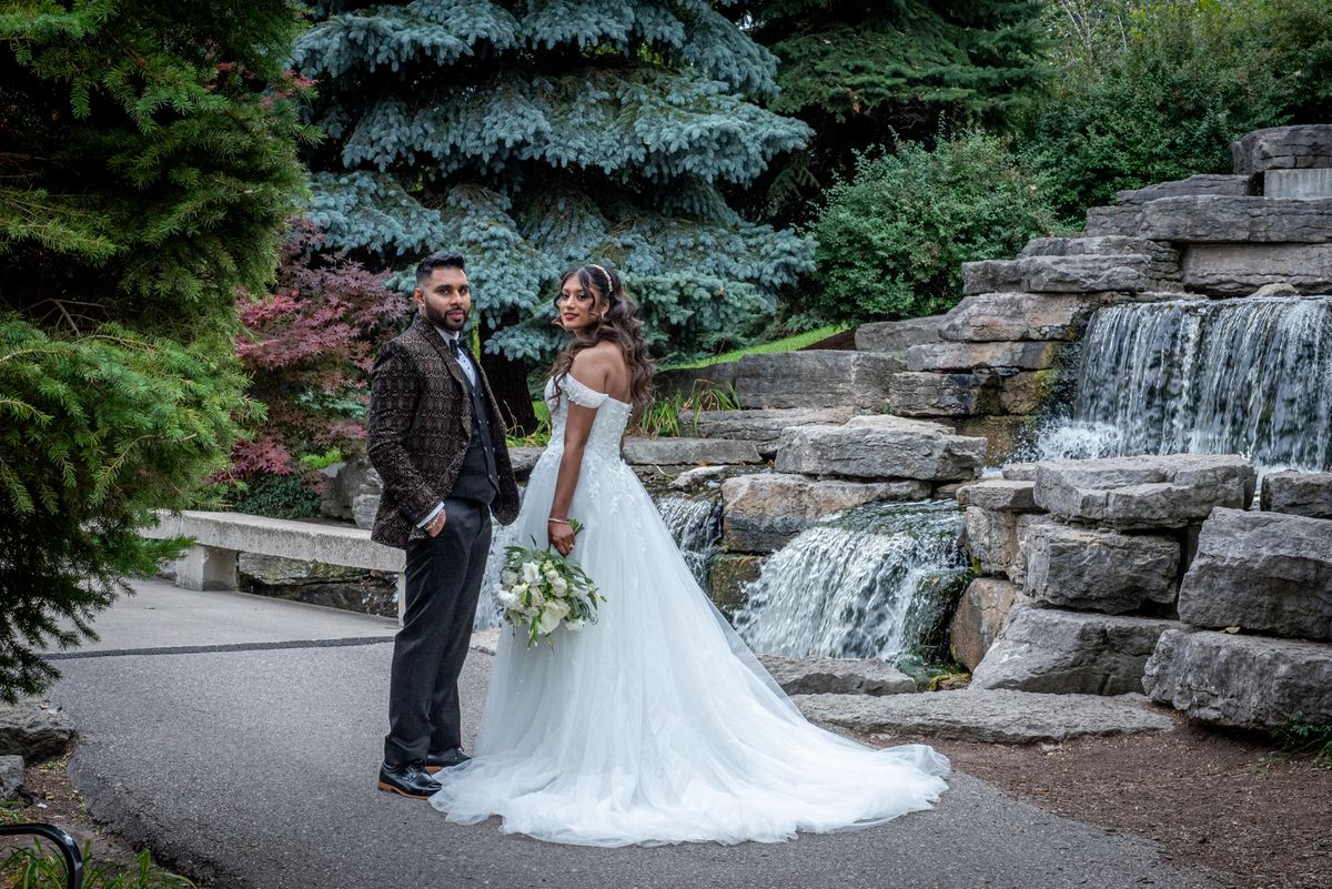 A bride in a white gown and groom by a stone waterfall in a garden, both smiling at the camera under diffused daylight.
