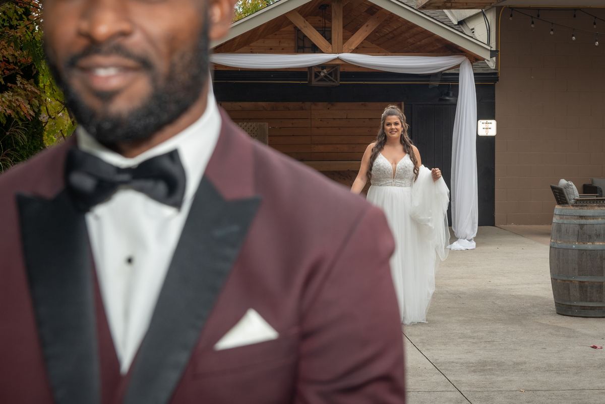 A bride smiles as she approaches her groom in a burgundy tuxedo, framed by out-of-focus fabric and soft lighting.
