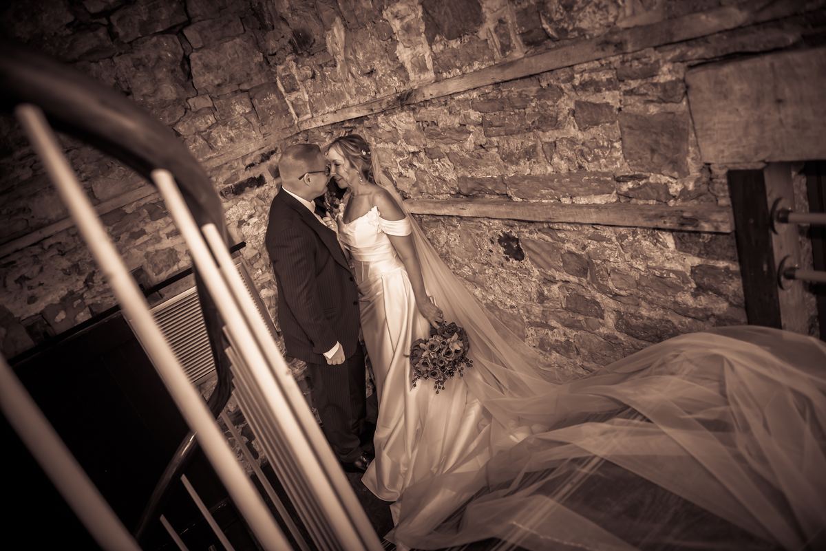 A newlywed couple leans in for a kiss on a stone staircase, the bride's veil flowing dramatically across the steps.