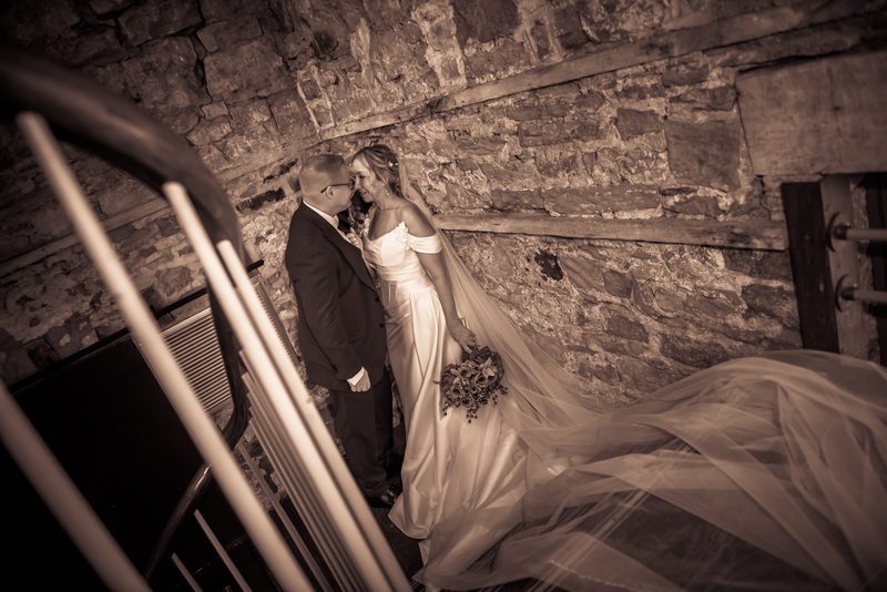 A newlywed couple leans in for a kiss on a stone staircase, the bride's veil flowing dramatically across the steps.