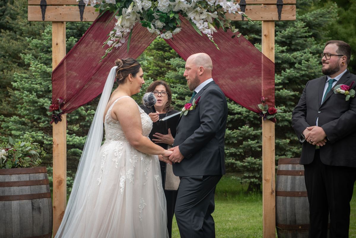 Bride and groom exchange vows under wooden arch adorned with burgundy fabric and white flowers, framed by evergreens.