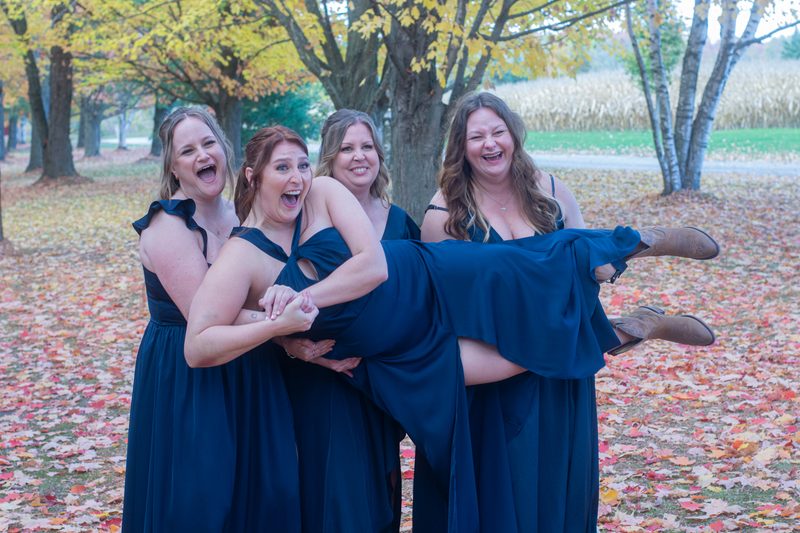 Four bridesmaids in navy gowns laugh as one is carried horizontally among autumn leaves under golden maple trees.