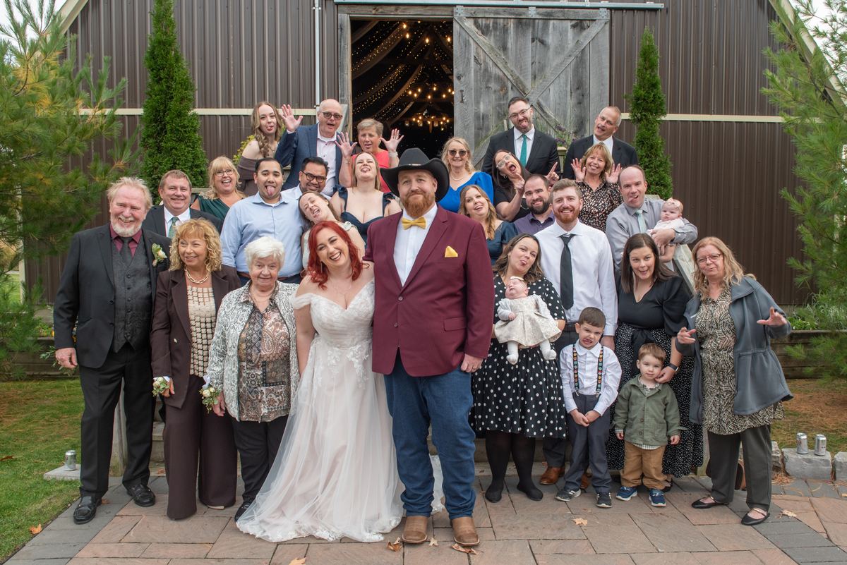 A wedding party outside a barn with string lights, the bride and groom at center surrounded by playful family and friends.