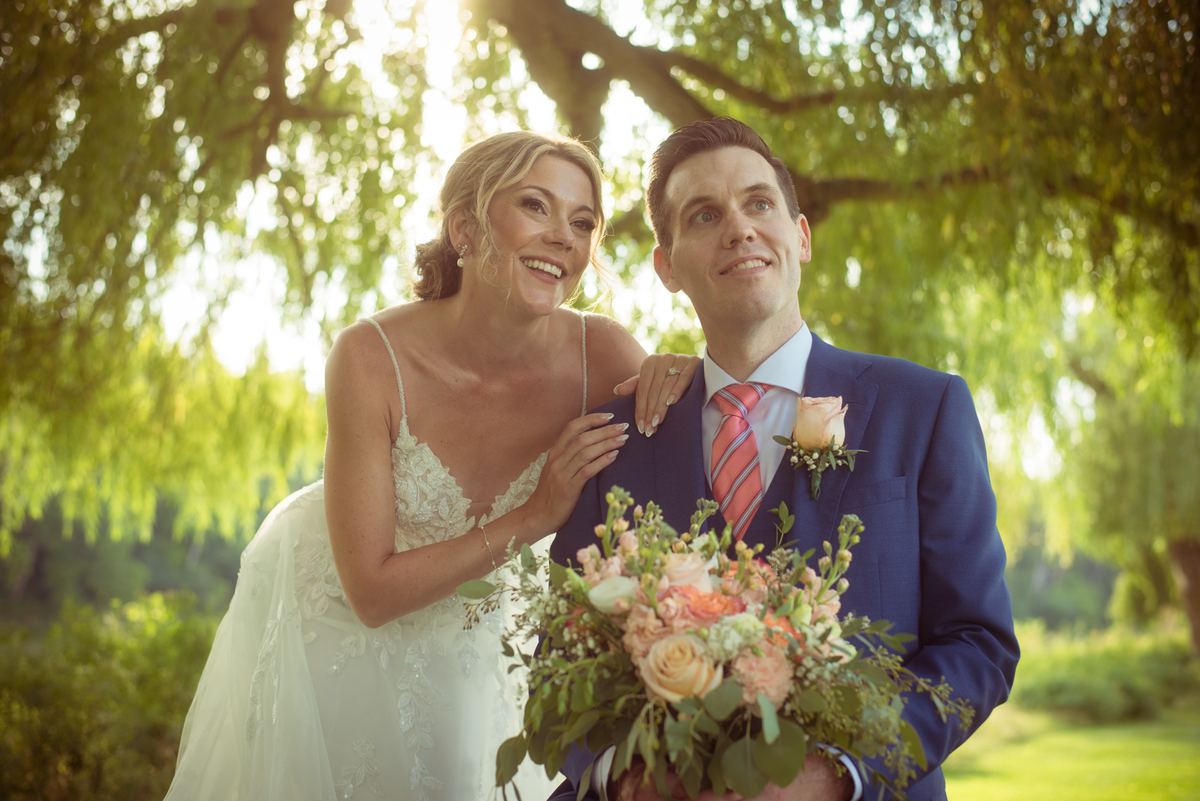 A smiling bride in a white lace gown leans against her groom beneath trees, both joyfully embracing the moment.