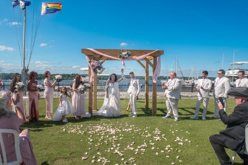 Bride and groom raise hands beneath wooden arch adorned with pink ribbons and flowers during sunny waterfront ceremony.