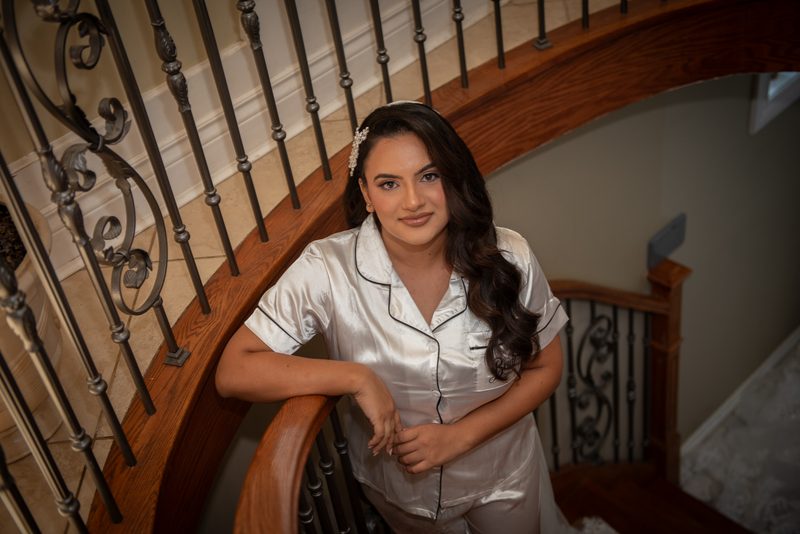 A bride in satin pajamas stands on a wooden staircase, smiling at the camera as warm light highlights the ornate railings.
