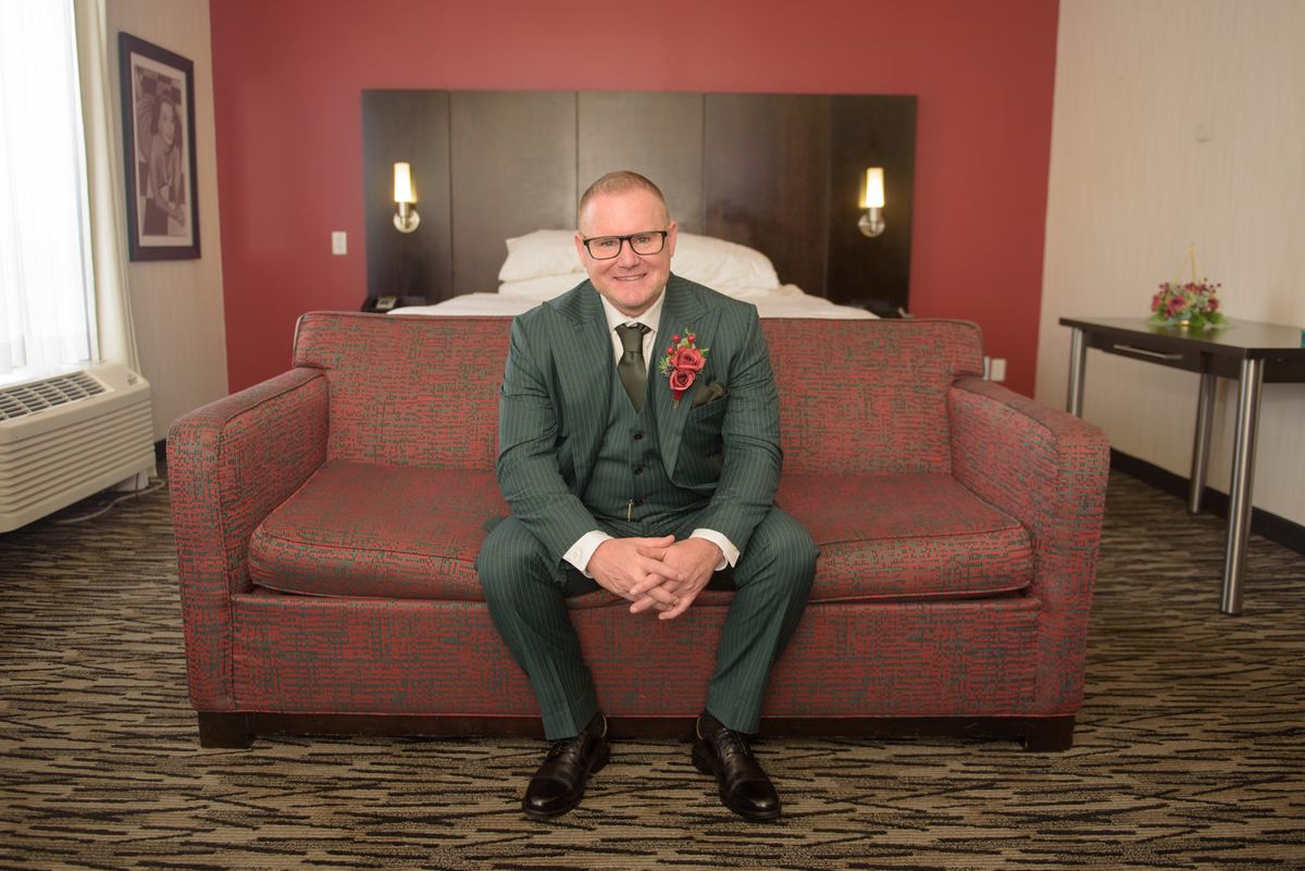 A groom in a green pinstripe suit sits smiling on a red patterned sofa in a hotel room, with a bed and window visible.