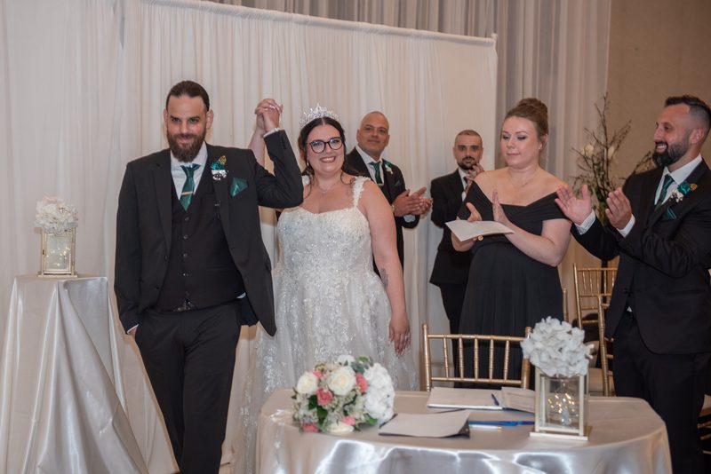 A smiling bride and groom in formal attire walk together as guests applaud, indoors with elegant white drapes.