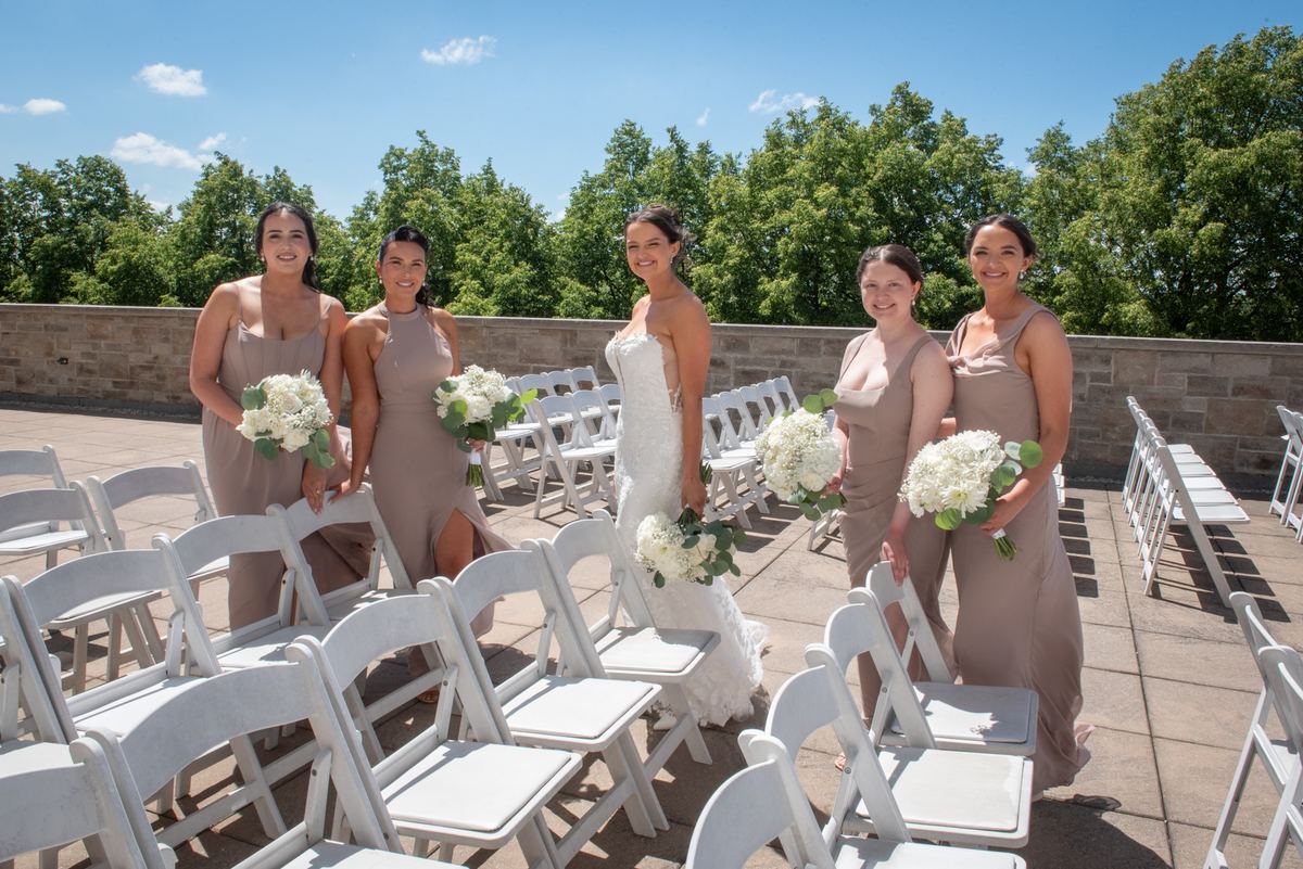 A bride and four bridesmaids smile with white floral bouquets outdoors against green trees and a stone wall.