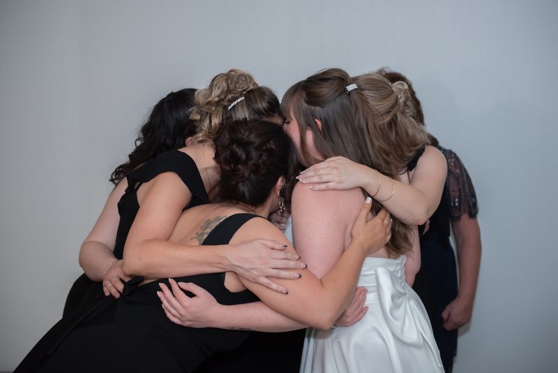 Women in a close embrace, including a bride in white, celebrate together warmly against a plain wall.