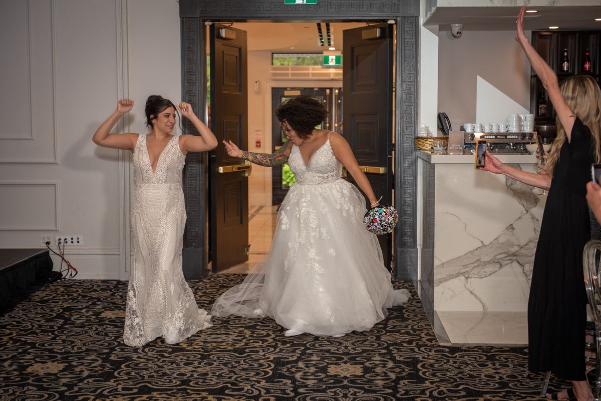 Two brides enter, one in a fitted lace gown with arms raised, the other in a voluminous tulle gown holding flowers.