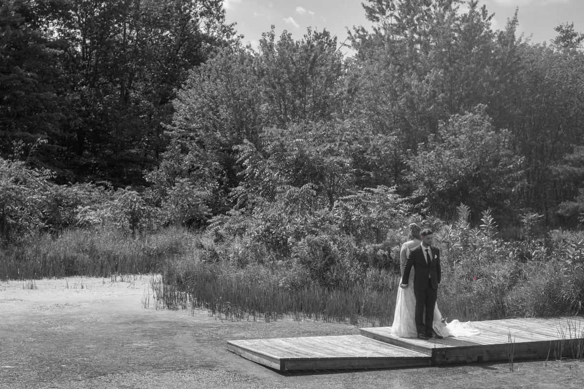 Darylleigh and CJ stand on a wooden platform near a marsh, surrounded by dense trees and reeds, in an intimate embrace.